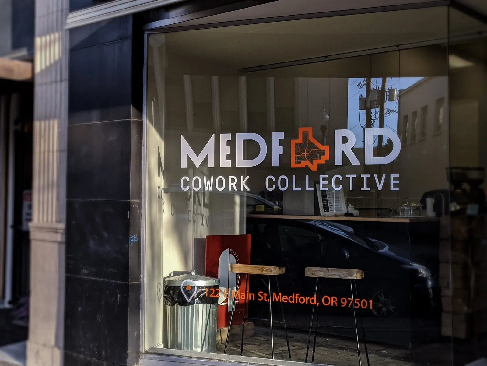 Storefront window with 'Medford Cowork Collective' lettering and logo, bar stools and the street reflected in the glass