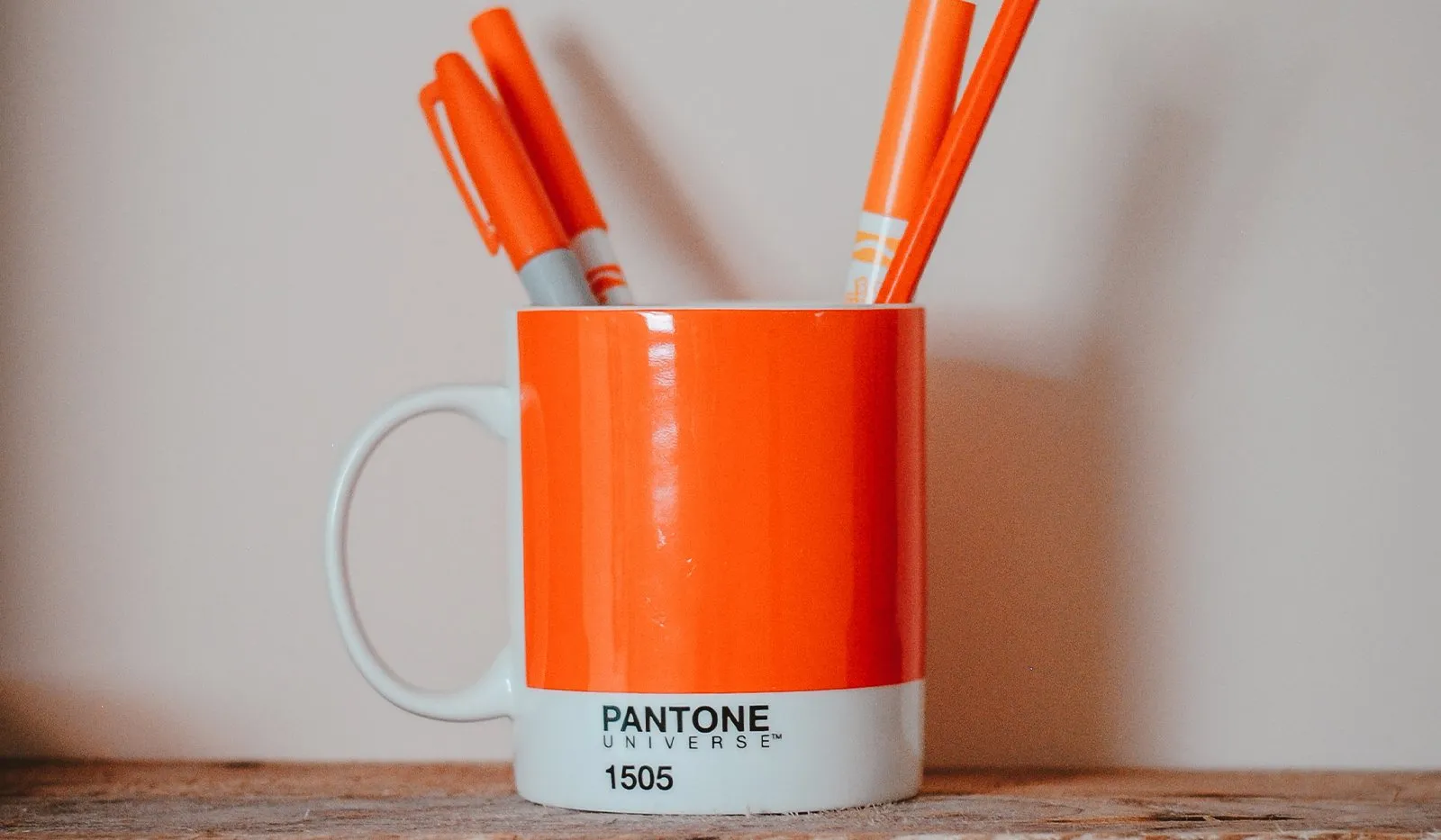 An orange Pantone 1505 mug holding orange marker pens on a wooden surface against a pale wall