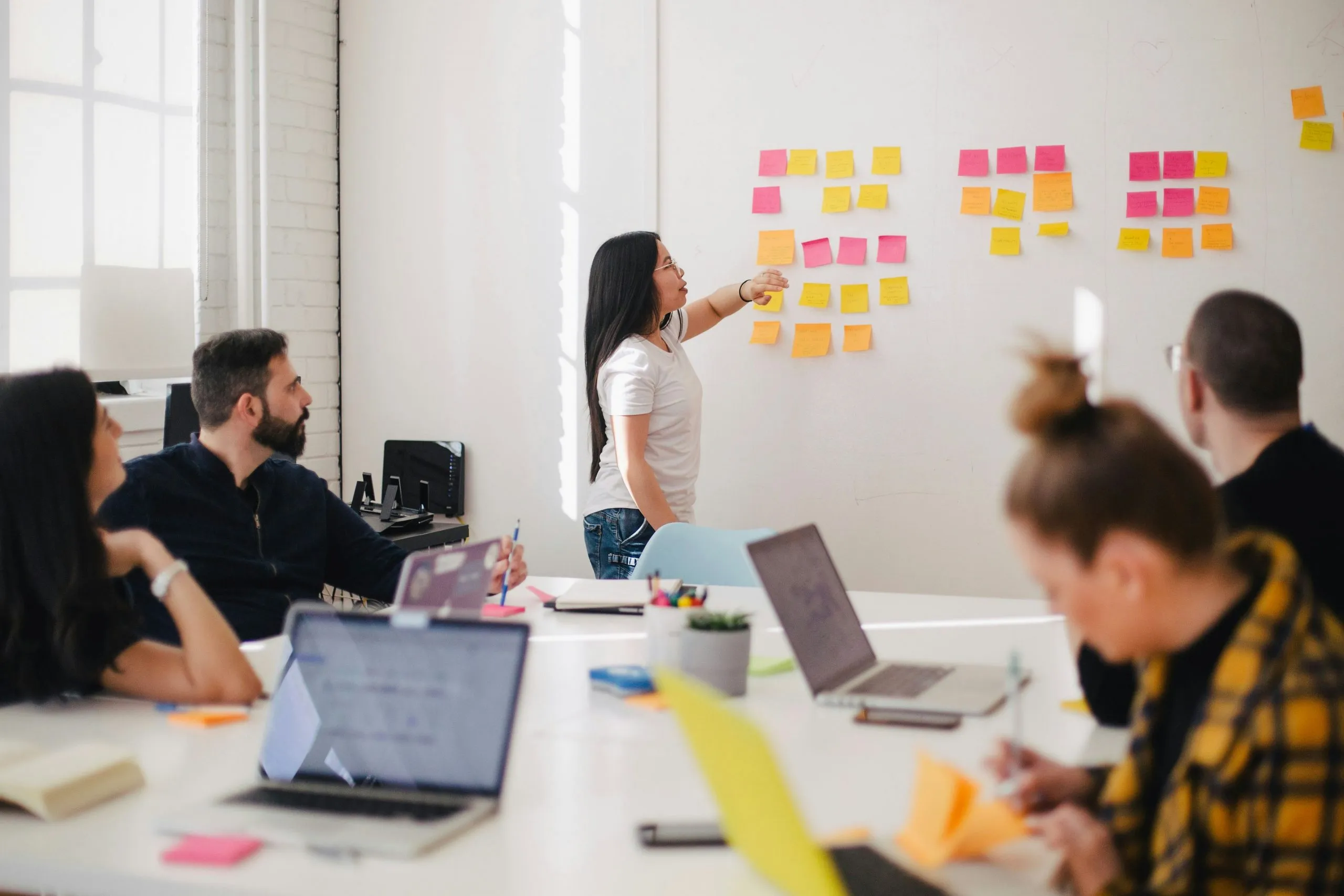 A woman arranges pink and yellow sticky notes on a white wall as seated colleagues with laptops watch in a bright office