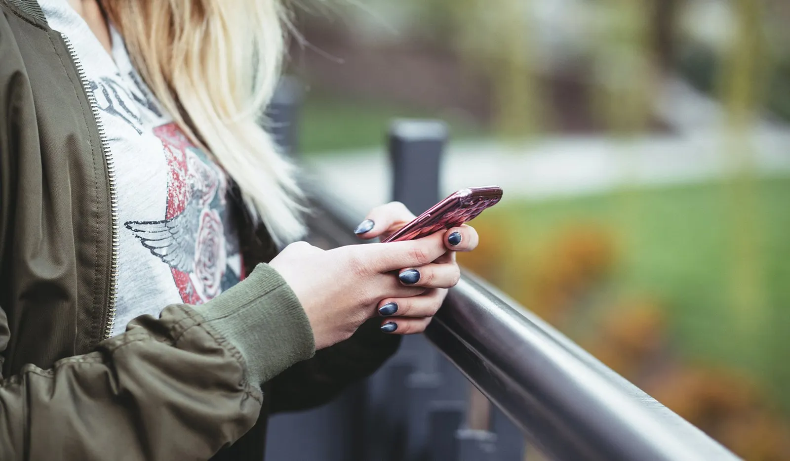 Close-up of a blonde woman in an olive bomber jacket texting on a red smartphone while leaning on a metal railing outdoors