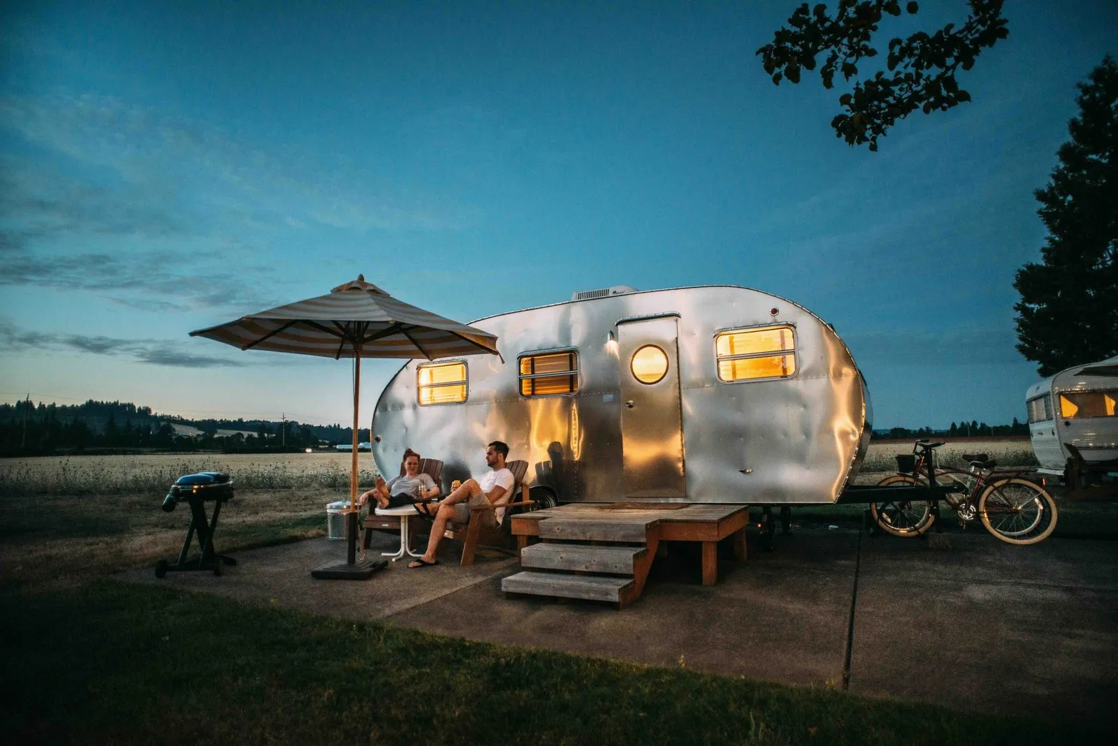 A vintage silver travel trailer glowing warmly at dusk, with two people relaxing under a patio umbrella beside an open field