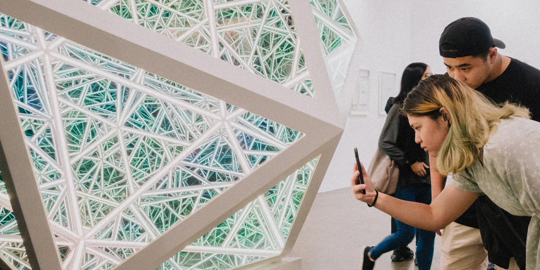 Two visitors photographing a large glowing geometric light sculpture inside a bright white museum gallery