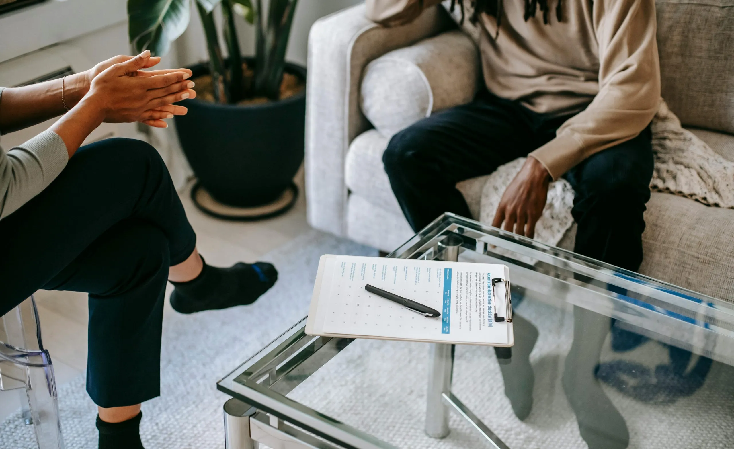 Therapy session with a client seated on a couch and a counselor across a glass table holding an assessment clipboard