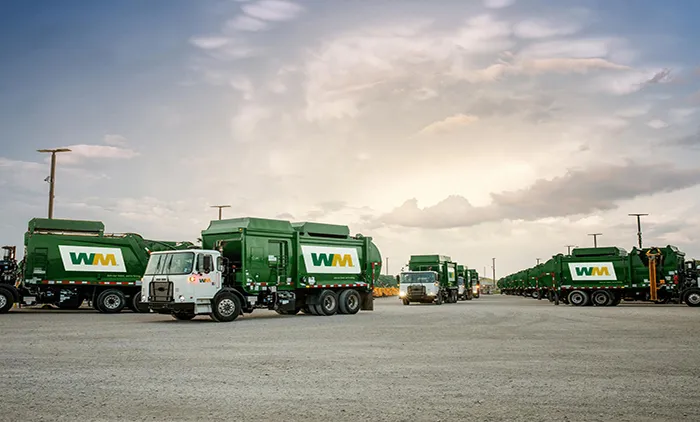 Fleet of green WM garbage trucks lined up across a paved lot under a cloudy sky