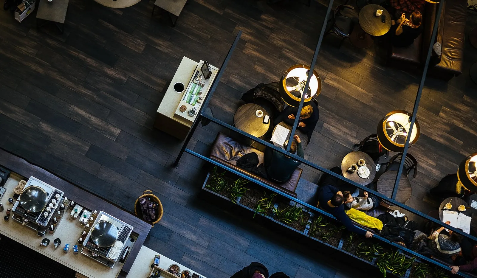 Overhead view of people seated at small round tables under hanging lamps in a dimly lit cafe with dark wood floors