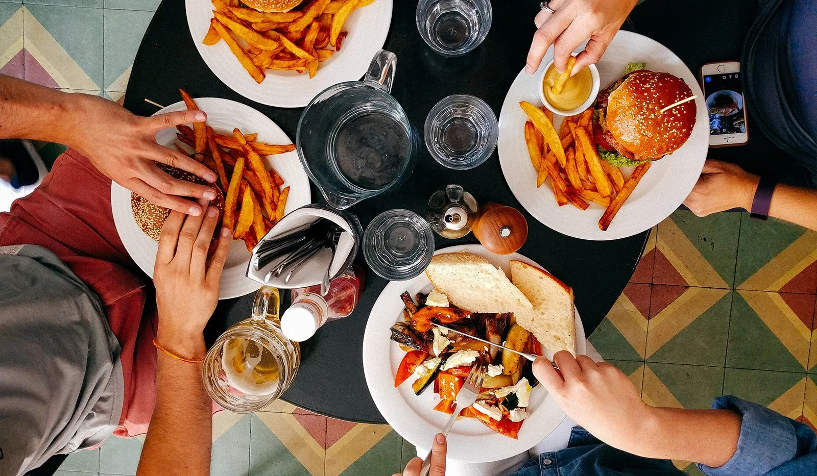 Overhead view of friends sharing burgers, fries and salad at a restaurant table on a colorful tiled floor