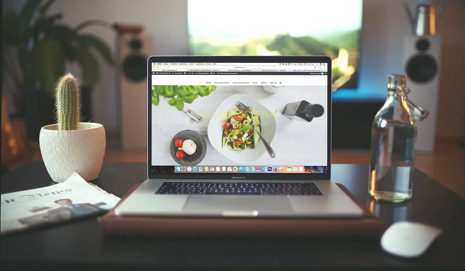 A MacBook on a desk showing a food website, beside a potted cactus, newspaper, mouse and glass water bottle