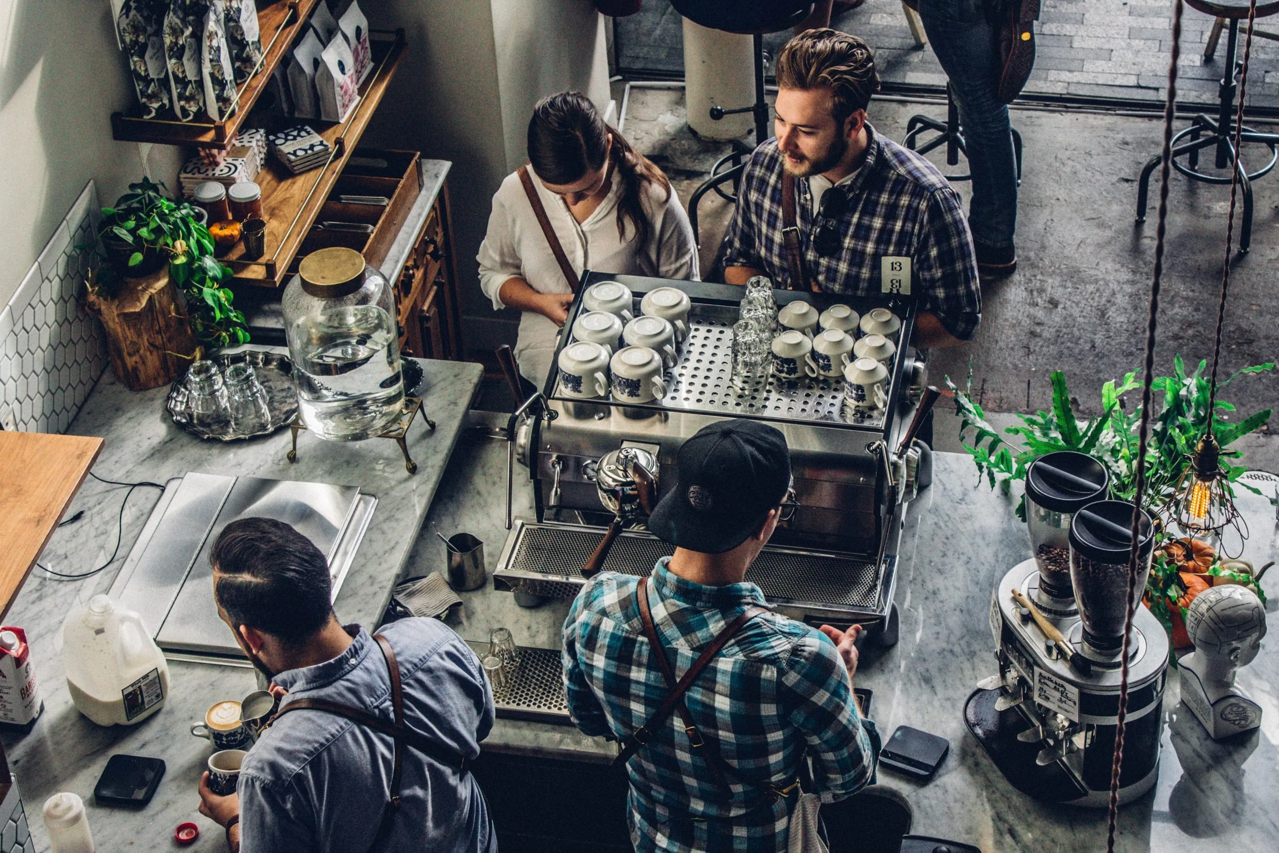 Overhead view of customers ordering at a cafe counter while baristas work behind an espresso machine stacked with cups