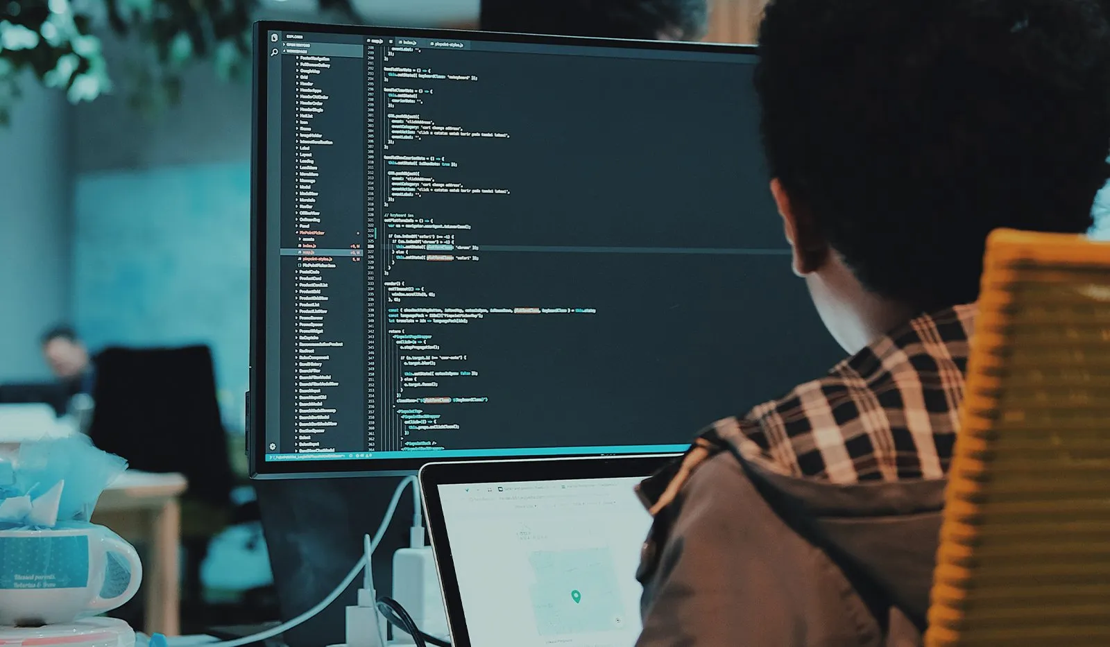 A developer in a plaid shirt seen from behind, working at a large monitor filled with code in a dim office