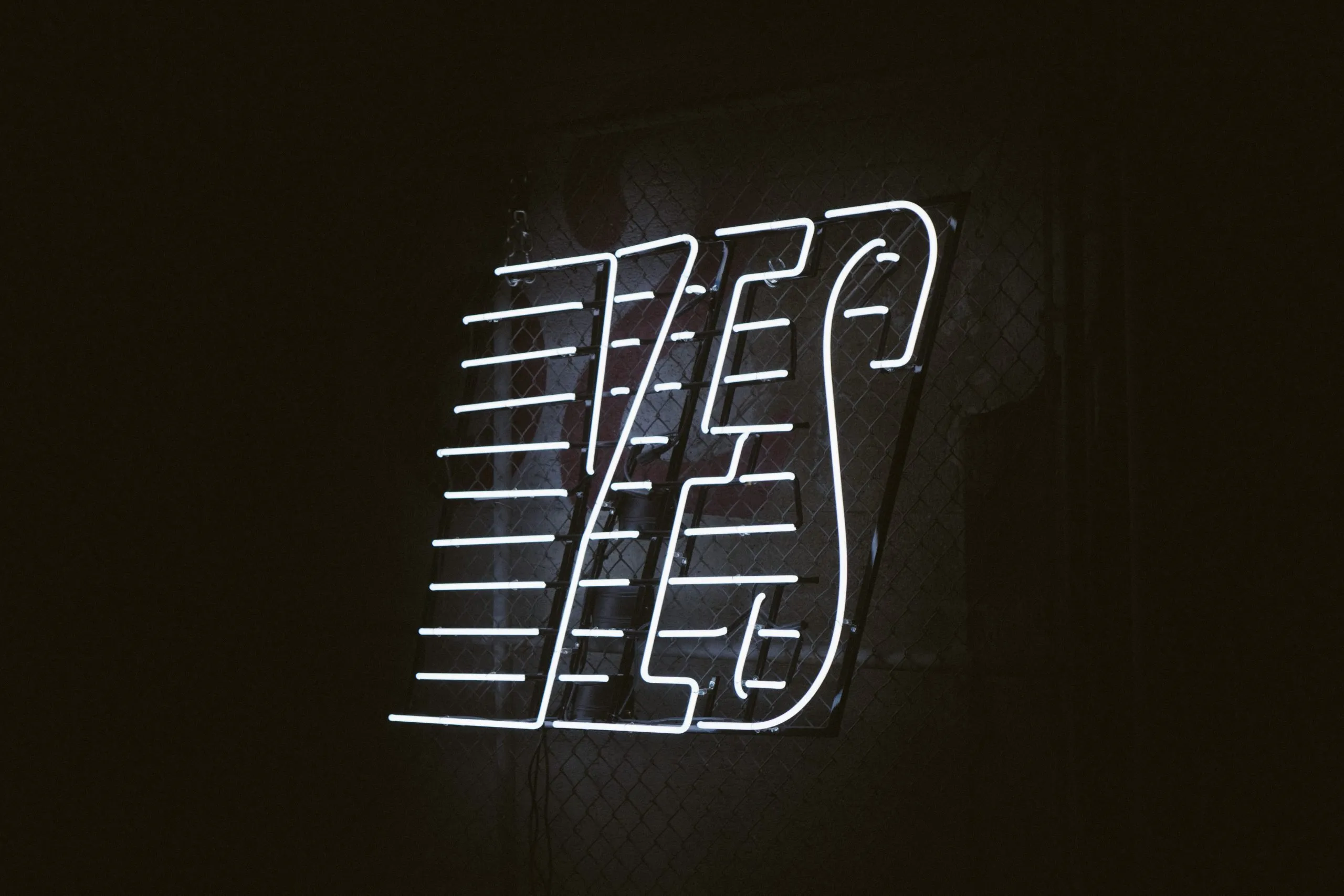 A glowing white neon sign spelling YES mounted on a chain-link fence in a dark room