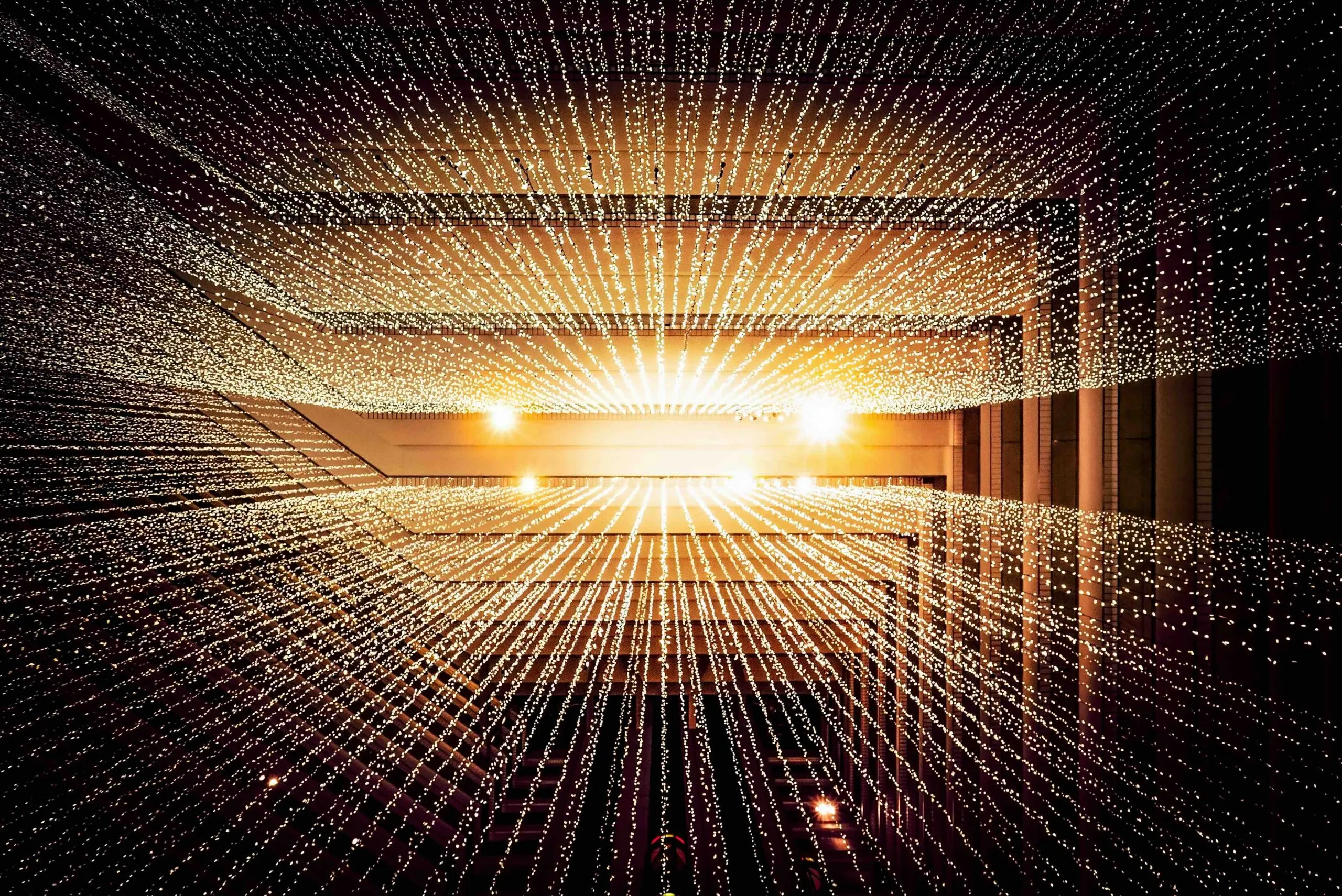 Looking up at a glowing grid of warm string lights stretching across a dark atrium ceiling toward two bright points