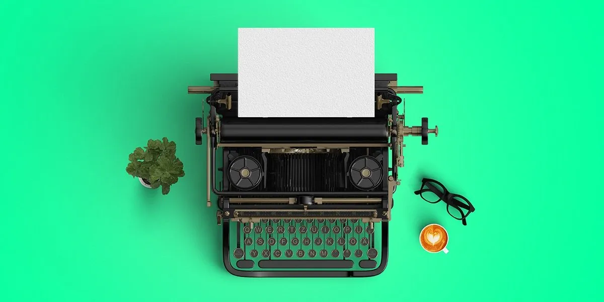 Overhead flat-lay of a vintage typewriter with blank paper, a potted plant, glasses and a heart-shaped latte on bright green