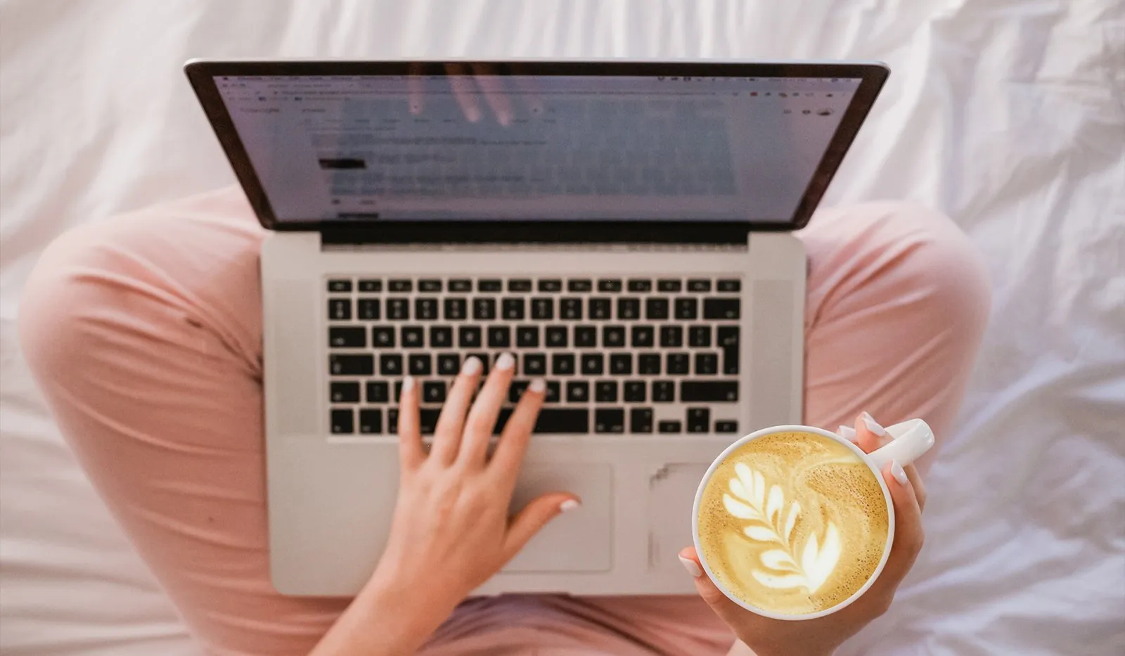 Overhead view of a person in pink pajamas typing on a laptop in bed while holding a cappuccino with leaf latte art