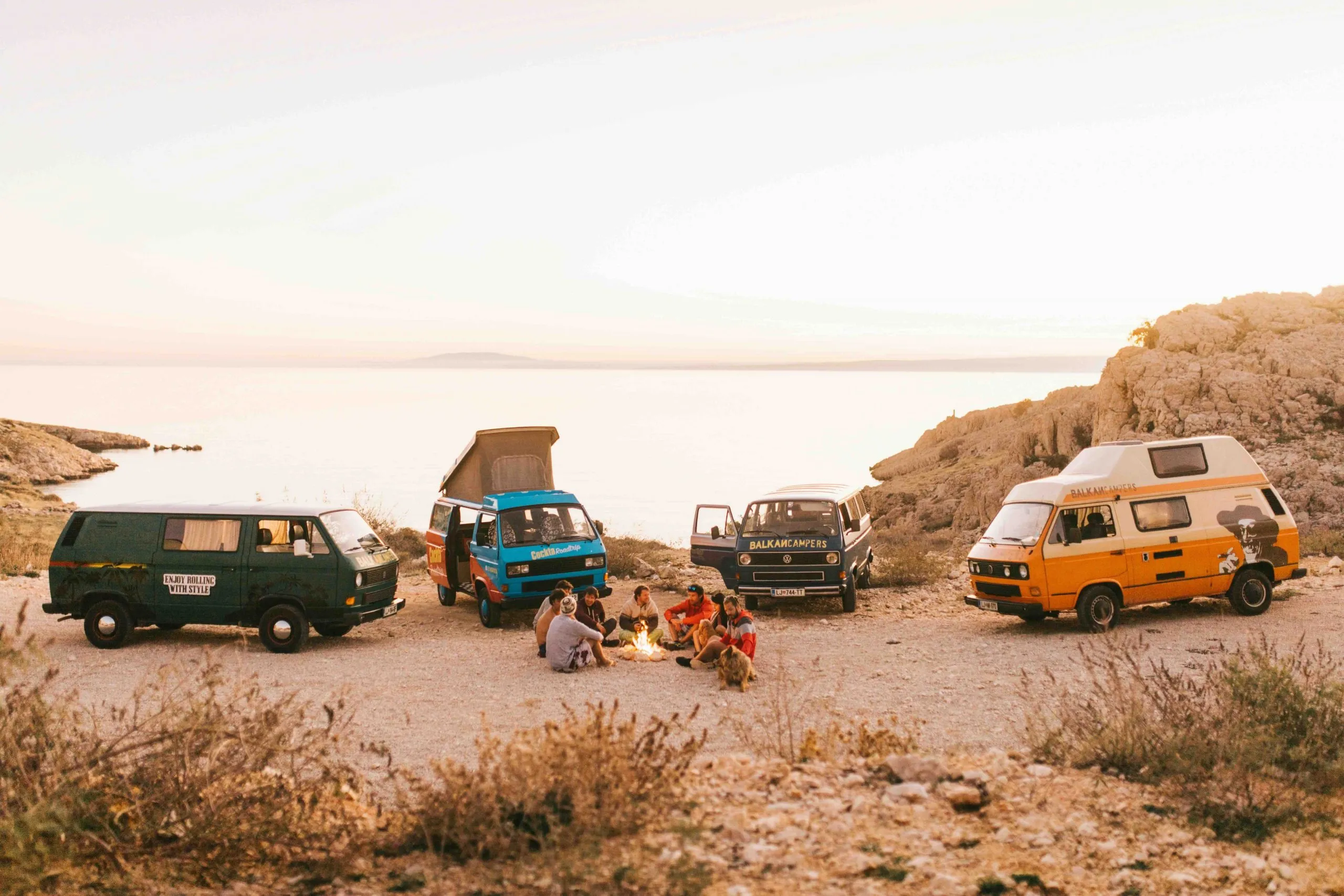Four camper vans parked on a rocky coastal cove at dusk as a group of friends sits around a small campfire