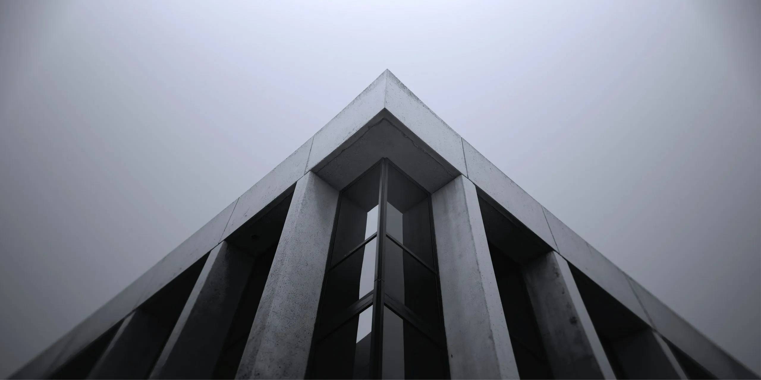 Upward view of the sharp concrete corner of a modern building with vertical fins and dark windows against a foggy sky