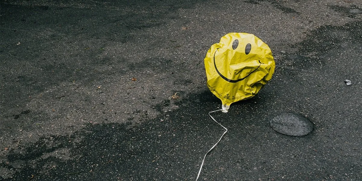 A deflated yellow smiley-face balloon lying on dark wet asphalt with its string trailing across the pavement