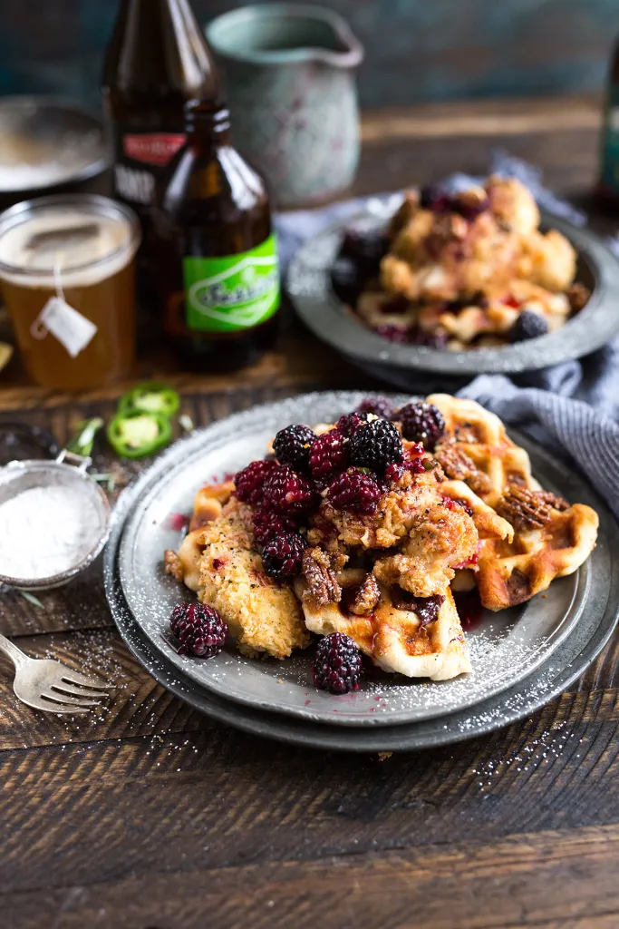 Plated waffles topped with blackberries and syrup on a dark table with bottles in the background