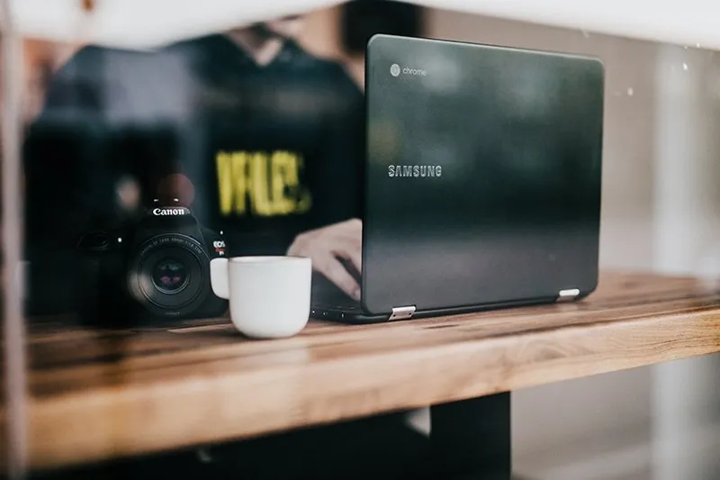 A Chromebook laptop and a DSLR camera beside a white mug on a wooden table