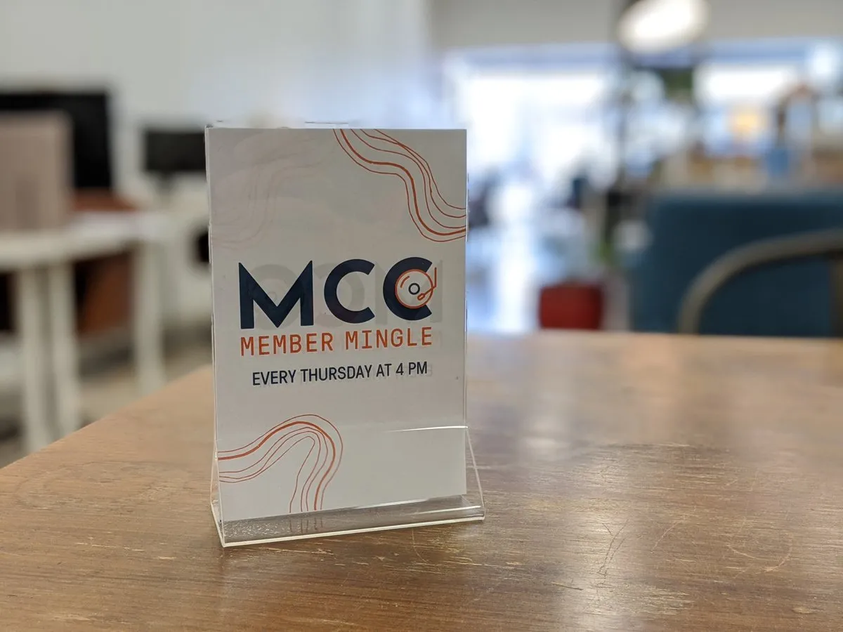 Branded table sign reading 'MCC Member Mingle, every Thursday at 4 PM' with orange map-line graphics